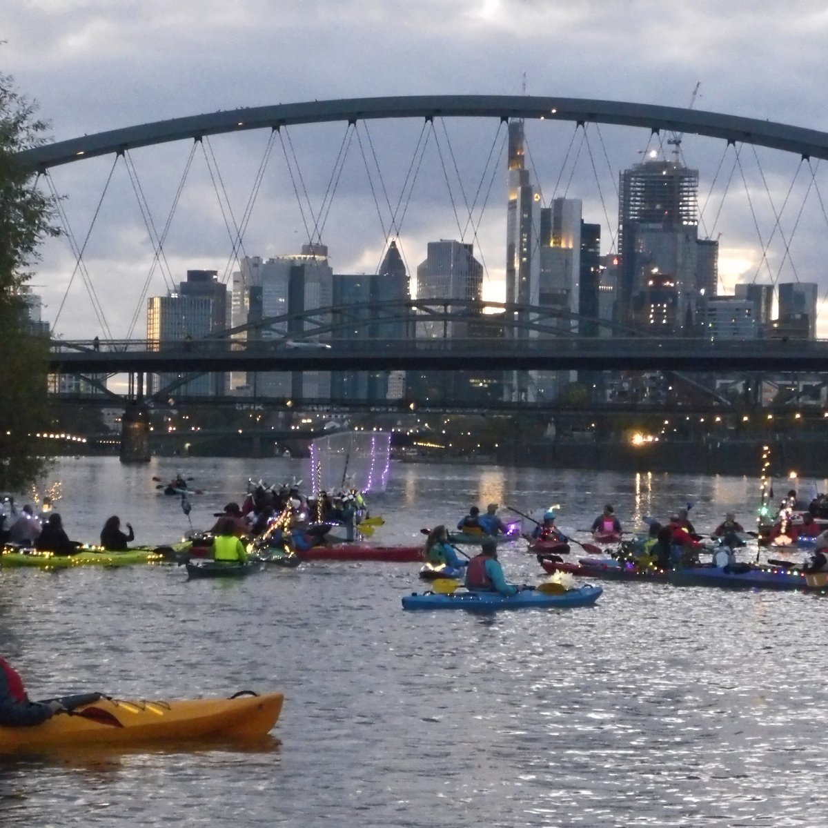 Langsam formieren sich die beleuchteten Boote vor der Fahrt auf dem Main. Im Hintergrund sieht man die Lichter der dunkler werdenden Innenstadt von Frankfurt